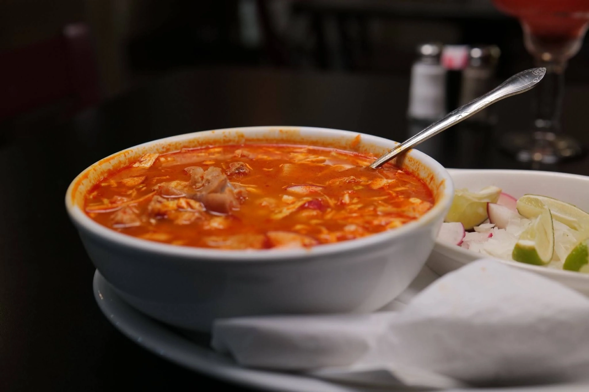 Bowl of red pozole with cabbage, radish, and lime garnishes on the side